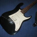 Černá elektrická kytara typu Stratocaster s bílým pickguardem a hnedým hmatníkem. Na kytaru jsou instalovány tři snímače typu single-coil a dva ovlád…