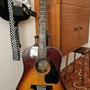 Akustická kytara s korpusem v přechodové barvě od žluté po tmavě hnědou a černým pickguardem. Na hlavě nástroje je bílý nápis Ibanez. Na spodní části…