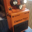 Oranžový efekt pedál s nápisy Distortion a DS-1. Na horní části jsou tři ovládací knoflíky, kontrolka s nápisem CHECK a přepínač s nápisem TONE. Na s…