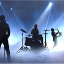 Fotografie zobrazuje siluety hudební kapely na scéně s modrým osvětlením a kouřem. Jsou vidět postavy kytaristů, bubeníka s bicí sadou a trumpetisty.…