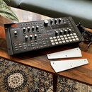Černý syntetizátor s nápisem HYDRASYNTH a logem Moog. Zařízení obsahuje zahlreiche ovládací knofíky, tlačítka a displej. Vedle přístroje leží dva kov…
