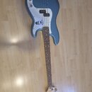 Modrá elektrická basová kytara s bílým pickguardem a hnedým hmatníkem. Na hlavě je viditelné logo Fender. Na pickguardu je nalepená bílá kulatá nálep…