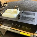 Černý mlhový generátor s bílou plastovou nádrží na tekutinu. Na boku přístroje je bílý nápis F-100 PERFORMANCE FOG GENERATOR a dvě varovné žluté znač…