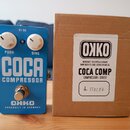 Modrý efekt pedál s nápisy COCA COMPRESSOR a OKKO HANDBUILT IN GERMANY. Na horní straně jsou dva bílé ovládací knoflíky s popisky PUSH a SING a vstup…