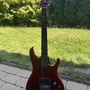 Elektrická kytara s červeným lesklým povrchem korpusu a hnědým hnedým hmatníkem. Na hlavě je bílý nápis Ibanez. Nástroj má dva humbuckery, kobylku s…