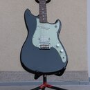 Černá elektrická kytara typu Stratocaster s bílým pickguardem a třemi snímači. Nástroj je umístěn na černém stojanu s červenými detaily. Povrch kytar…