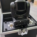 Černý movable head reflektor umístěný v transportním flightcasu s hliníkovými hranami. Na zadním panelu zařízení jsou nápisy DMX IN, DMX OUT a logo s…