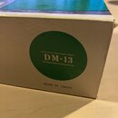 Kartonová krabice s modrým vrchem a bílými boky. Na boku je zelený kruh s nápisem DM-13 a pod ním text MADE IN JAPAN. Hrany krabice vykazují mírné op…