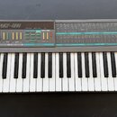 Šedý syntetizátor s klaviaturou a ovládacím panelem. Na horní části je nápis KORG POLY-800, digitální displej a řada tlačítek s čísly od 11 do 48. Pa…