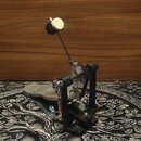 Černý bubnový pedál s kovovými mechanismy a stříbrným šlapadlem. Konstrukce obsahuje řetězový pohon a nastavitelné pružiny. Na povrchu nejsou vidět ž…