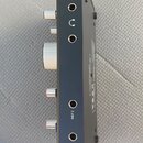 Černý elektronický přístroj s kovovým šasi a stříbrnými ovládacími prvky. Na boku jsou čtyři jack konektory, u dvou z nich jsou nápisy INST 1 a INST…