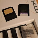 Bílý klávesový syntetizátor s nápisy YAMAHA, MUSIC SYNTHESIZER a MX61. Na přístroji leží dvě černé paměťové karty, z nichž jedna má nápis OLYMPUS a S…