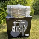 Na krabici s nápisy SONOR, KOMPRESSOR SNARE DRUM SERIES a sonor.com stojí zabalený malý buben. Nástroj je stříbrný s kovovým povrchem a je celý obale…