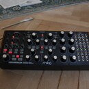 Černý analogový polyrytmický syntetizátor s nápisy SUBHARMONICON a moog. Přístroj obsahuje řadu bílých otočných ovladačů, červených podsvícených tlač…