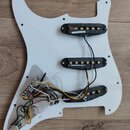 Bílá plastová pickguard pro elektrickou kytaru s třemi instalovanými snímači. Snímače mají černé plastové kryty a viditelné mosazné póly, z nichž něk…