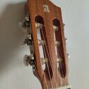 Detail hlavy akustické kytary z hnědého dřeva s bílým logem ve tvaru stylizovaného písmene A. Na hlavě jsou vidět tři kovové ladičky s bílými plastov…
