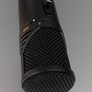 Černý kondenzátorový mikrofon s kovovou síťkou. Na těle je zlatý nápis -10dB a v horní části jsou dva ovládací knoflíky a tlačítko s nápisem MUTE. Na…