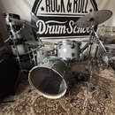 Šedo-stříbrná bicí souprava s chromovaným hardwarem umístěná na vzorovaném koberci. V pozadí je vidět černobílý banner s nápisem ROCK & ROLL Drum Sch…