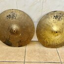 Dvě mosazné činely s nápisem MAGIC BRASS. Levý činele má v dolní části nápis HI-HAT TOP 14" a vykazuje výrazné oxidace a tmavé skvrny. Pravý činele m…
