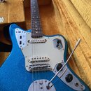 Modře třpytivá elektrická kytara s bílým pickguardem v hnědém polstrovaném kufru. Na kovovém krytu spodního tremola je nápis Fender. Nástroj má hnědý…