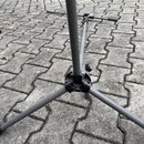 Šedý kovový stativ s třemi rozkládacími nohami umístěný na dlažbě. Na centrálním spoji je vidět černý plastový uzel s utahovacím šroubem. Povrch kovo…