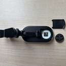 Černý plastový clip-on ladicí přístroj s odpojeným víčkem baterie. V prostoru pro baterii je viditelný kovový kontakt a vedle přístroje leží knoflíko…
