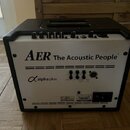 Černý kytarový kombozesilovač s bílým předním panelem. Na panelu je uveden nápis AER The Acoustic People® a model Alpha plus. V horní části jsou ovlá…