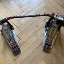 Dvojpedál na bicí buben v stříbrno-černé barvě s červenými detaily na spojovací tyči. Na obou pedálech je vidět nápis dw 9000 a text The Drummer's Ch…