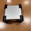Stříbrné kovové zařízení s čtyřmi černými gumovými nárožníky. Na zadní straně jsou dva konektory XLR s nápisy BAL a jeden menší vstup. Povrch je matn…