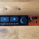 Detailní pohled na ovládací panel černého elektronického zařízení s oranžovými prvky. Na panelu jsou vidět modré tlačítka, velký černý otočný knoflík…
