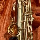 Detailní pohled na část zlatého saxofonu v hnědém polstrovaném pouzdře. Jsou vidět kovové mechanismy, klapy a spojovací tyče s lesklým povrchem. Na t…