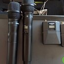 Dva černé bezdrátové mikrofony s mřížkami z kovového pletiva a dva odpovídající černé vysílače. Na těle levého mikrofonu je nápis SHURE a označení SM…