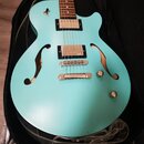 Světle modrá polodublá elektrická kytara s dvěma f-otvory v těle, umístěná v černém obalu. Nástroj má dva humbuckery a stříbrný hardware. Povrch těla…
