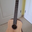 Akustická kytara s přírodním dřevěným povrchem korpusu a tmavým hmatníkem. Na hlavě je bílý nápis Cort. Nástroj stojí v černém držáku na světlé dlažb…