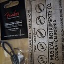 Fotografie zobrazuje černý klíčenka ve tvaru kytarového jacku v originálním balení s nápisy Fender, GUITAR PLUG KEYCHAIN a FREE ONLINE LESSONS. Vedle…