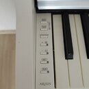 Detail ovládacího panelu bílého digitálního pianina. Na panelu jsou tlačítka s nápisy DEMO/SONG, PIANO/VOICE, METRONOME, + R, - L, REC a PLAY, vedle…