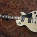 Bílá elektrická kytara typu Les Paul s černými snímači a ovládacími knoflíky. Na hlavě hmatníku je viditelné logo Epiphone. Nástroj vykazuje známky p…