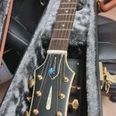 Akustická kytara v černém tvrdém kufru s šedým polstrováním. Na černé hlavě hmatníku je zlatý nápis Epiphone a bílý nápis MASTERBILT s malým modrým l…
