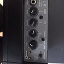 Černý kompaktní basový zesilovač s šedým ovládacím panelem. Na panelu jsou nápisy FLY, 2 WATT MINI AMP a BASS, čtyři černé otočné ovladače s popisky…