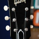 Fotografie zachycuje hlavu černé kytary s bílými ladičkami. V horní části je umístěno zlaté logo Gibson. Na čelní straně je vidět bílý ornament ve tv…