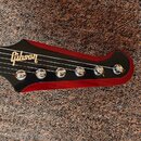 Fotografie zachycuje hlavu a část hmatníku elektrické kytary. Černý povrch hlavy nese bílé logo Gibson, zatímco okraje jsou provedeny v červeném dřev…