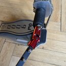 Detailní pohled na část mechanismu basového pedálu. Na šedém povrchu je černý nápis „DW 9000“. Součástí konstrukce je červený anodovaný kovový spoj s…