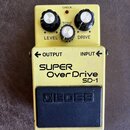 Žlutý kytarový efekt pedál značky BOSS, model SUPER Over Drive SD-1. Na horní části jsou tři černé ovládací knoflíky s bílými vršky a nápisy LEVEL, T…