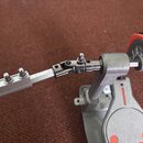 Detailní pohled na kovový mechanismus basového pedálu v stříbrné barvě. Je vidět spojovací tyč s několika šrouby a červený prvek u úderníku. Povrch k…