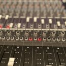 Detailní pohled na ovládací panel míchacího pultu s řadou vertikálních faderů a rotačních potenciometrů. Na panelu jsou vidět bílá tlačítka s nápisy…