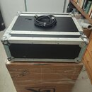 Černý transportní flightcase s hliníkovými hranami a kovovými rohy. Na horním víku leží smotaný černý kabel. Case je umístěn na kartonové krabici s n…