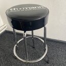 Černý kulatý taburet s čalouněným sedákem a chromovanou kovovou konstrukcí s opěrkou na nohy. Na horní části sedáku je bílý nápis th.mann MUSIC IS OU…