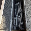 Otevřený hliníkový transportní kufr s vnitřním polstrováním z šedé pěny. Uvnitř se nachází několik černých kovových stativů v různém rozkladu. Na pov…