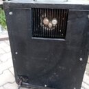 Zadní strana černého komba s rukojetí z plastu a kovu. V horní části je umístěna ventilační mřížka, za kterou jsou vidět tři elektronky. Povrch je zm…