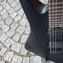 Detailní pohled na horní roh černého trupu elektrické kytary s matným povrchem. Je vidět část hmatníku s pražci, struny a černý most. Nástroj je polo…